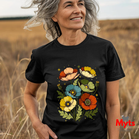 A smiling older woman standing in a field of dry grass wearing a black short-sleeve T-shirt with a colorful floral poppy print on the front. The brand name 'Myts' appears in red in the bottom right corner.