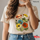 A woman wearing a beige short-sleeve t-shirt with a colorful poppy floral graphic on the front, paired with blue jeans. The image features a mirror-selfie style with a phone partially covering the face, and text overlays reading 'Natural' and 'Myts designs'.