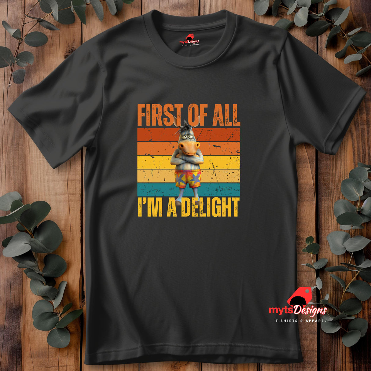 Casual black tee displaying a retro-style donkey illustration with bold “First of All, I’m a Delight” vintage text, flat lay on wooden boards