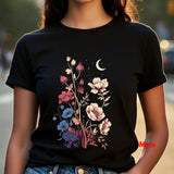 A woman wearing a black short-sleeve t-shirt with a colorful graphic of wildflowers, a crescent moon, and stars on the front, set against a blurred outdoor background.