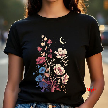 A woman wearing a black short-sleeve t-shirt with a colorful graphic of wildflowers, a crescent moon, and stars on the front, set against a blurred outdoor background.
