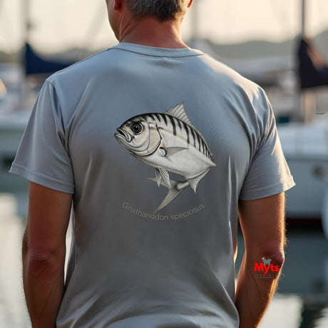 Man wearing a t-shirt with a fish design on the back, standing by water with boats in the background.