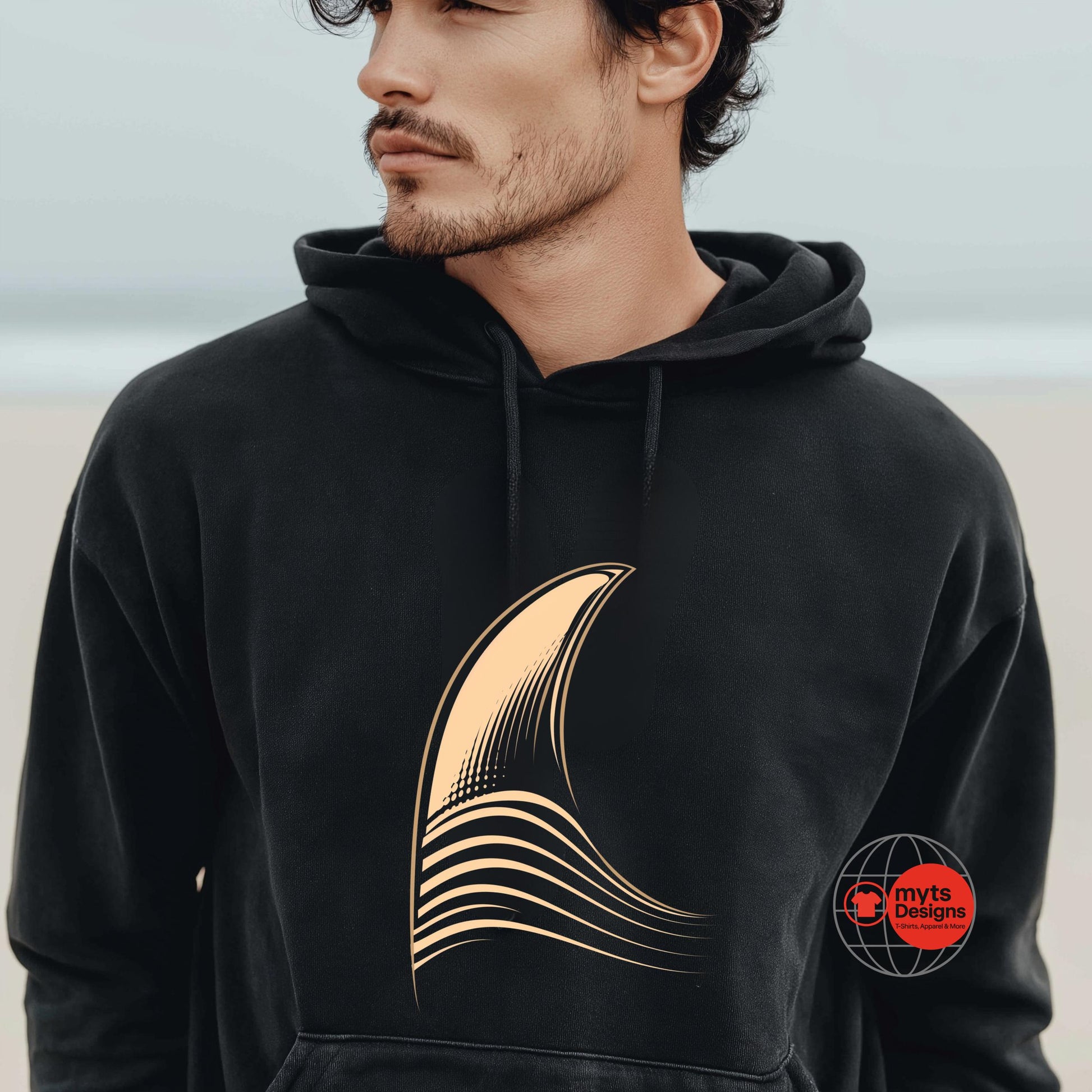 Man wearing a black hoodie with a gold shark fin design on a beach