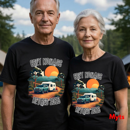 An older man and woman standing outdoors wearing matching black graphic T-shirts. The shirts feature a sunset landscape print with the text 'GREY NOMADS RETIRED 2025' and an illustration of a blue pickup truck and white camper.