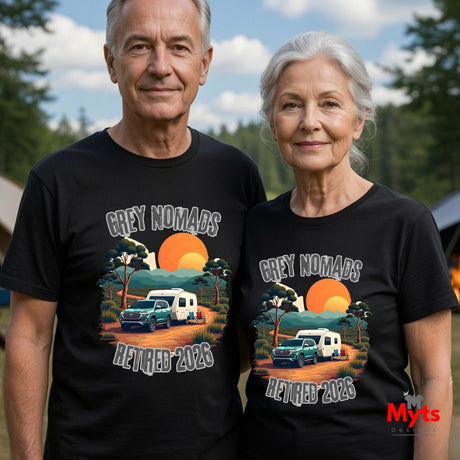 An older man and woman standing outdoors wearing matching black graphic T-shirts. The shirts feature a sunset landscape print with the text 'GREY NOMADS RETIRED 2025' and an illustration of a blue pickup truck and white camper.
