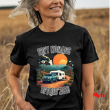 A woman wearing a black graphic t-shirt in an outdoor field. The t-shirt features a colorful retro-style illustration of a green ute and a white caravan on an orange dirt road, with the text 'GREY NOMADS RETIRED 2026' in a distressed white font.