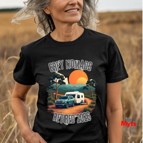 A woman wearing a black graphic t-shirt in an outdoor field. The t-shirt features a colorful retro-style illustration of a green ute and a white caravan on an orange dirt road, with the text 'GREY NOMADS RETIRED 2026' in a distressed white font.