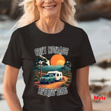 A woman wearing a black graphic t-shirt with a retro camping scene that reads 'GREY NOMADS RETIRED 2026'. The background is a blurred beach setting.