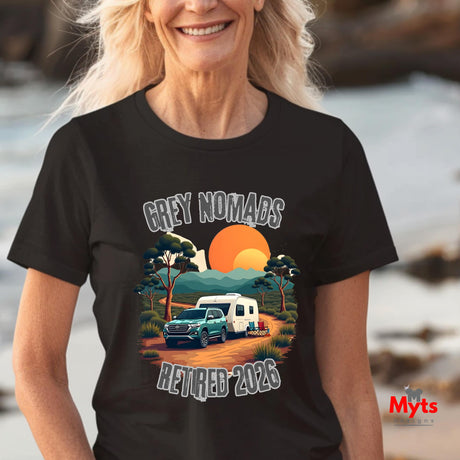 A woman wearing a black graphic t-shirt with a retro camping scene that reads 'GREY NOMADS RETIRED 2026'. The background is a blurred beach setting.
