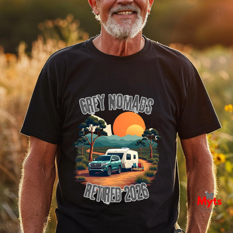 A smiling older man wearing a black graphic t-shirt in an outdoor field at sunset. The t-shirt features a colorful graphic of a green truck and a white caravan under a large orange sun, with the text 'GREY NOMADS RETIRED 2026' in a stylized font.
