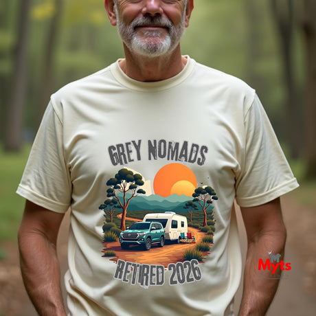 A man wearing a cream-colored graphic t-shirt in an outdoor forest setting. The t-shirt features a retro-style illustration of a green pickup truck towing a white camper trailer on a dirt road, with a large orange sun and the text 'GREY NOMADS RETIRED 2026'.