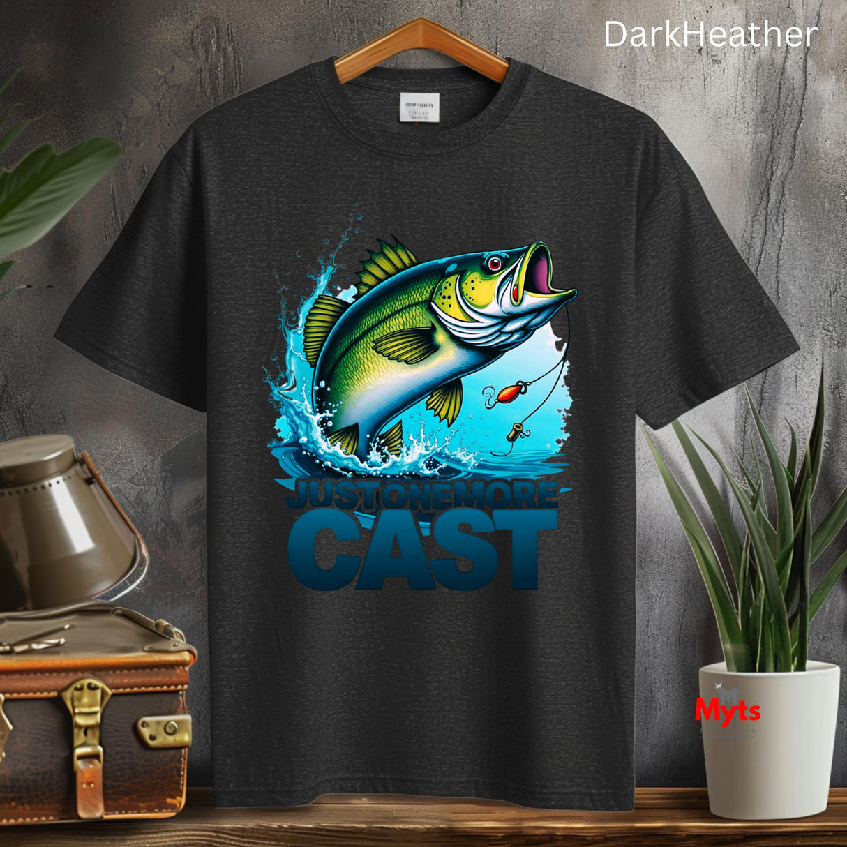 Black t-shirt with a fish graphic and 'CAST' text on a wooden surface with plants and a suitcase.