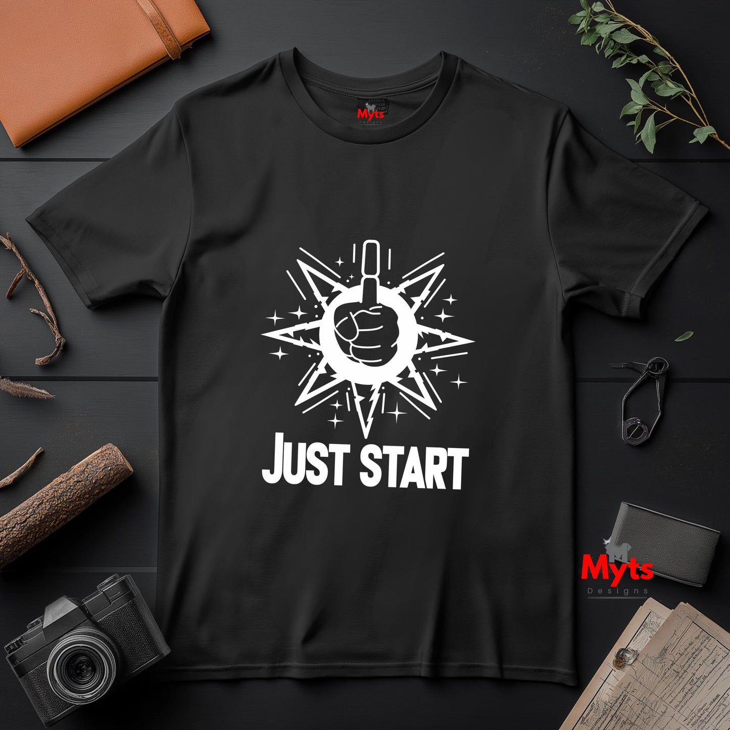 Black t-shirt with white graphic and 'JUST START' text on a dark surface with camera and books.