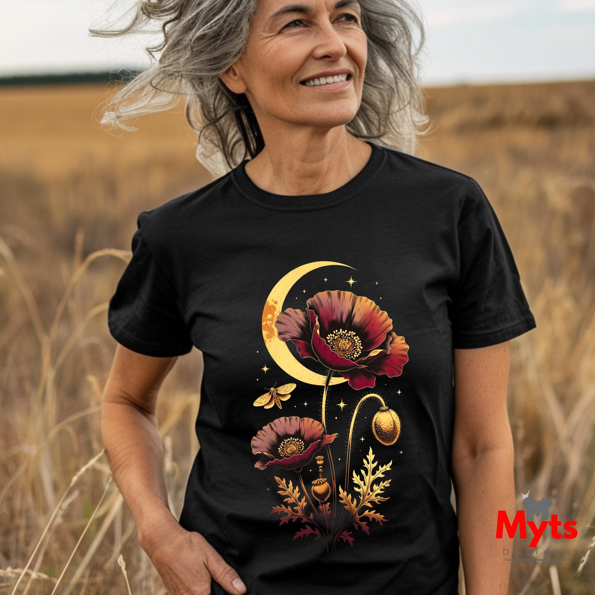 A smiling older woman standing in a field of dry grass wearing a black graphic T-shirt. The shirt features a detailed print of red poppy flowers, a crescent moon, stars, and small butterflies in warm gold, orange, and red tones.