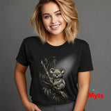 A smiling blonde woman wearing a black short-sleeve graphic t-shirt with a detailed illustration of a koala on a tree branch, set against a solid gray background.