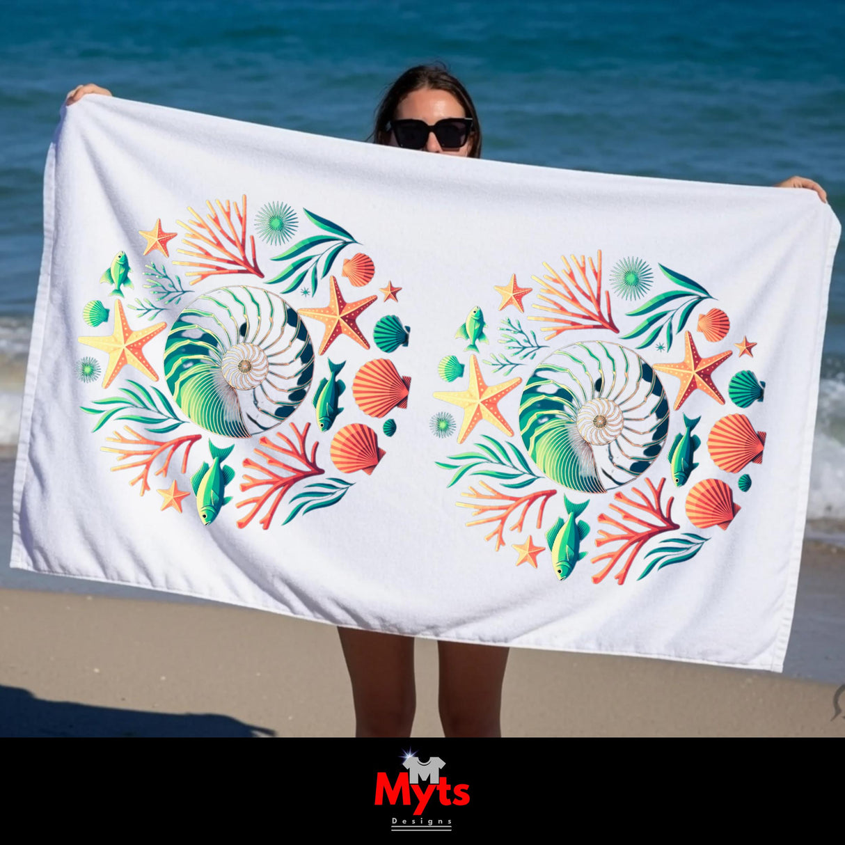 A white beach towel with a colorful nautical print of seashells, starfish, coral, and fish, held up by a person on a beach with the ocean in the background.