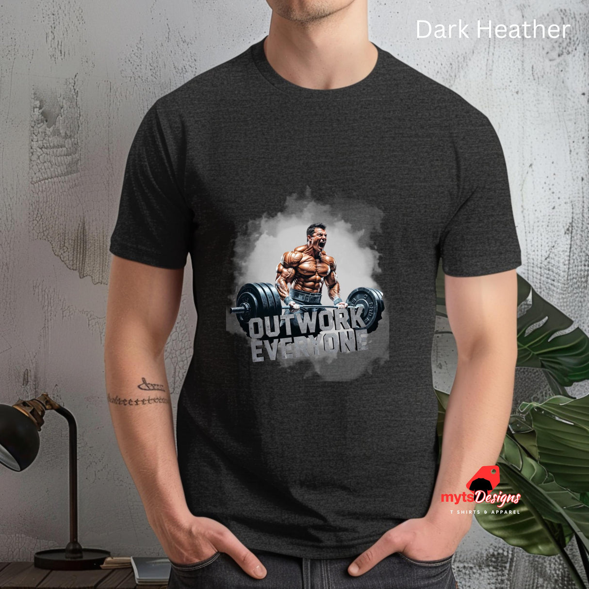 Person wearing a dark heather t-shirt with a graphic design and text 'Outwork Everyone' on a textured wall background. heather