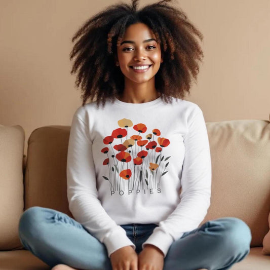 A woman sitting cross-legged on a couch wearing a white sweatshirt featuring a minimalist poppy flower design. The graphic displays red, orange, and muted gold poppies with slender stems and leaves, and the word “POPPiES” printed beneath. The cozy, casual outfit has a soft, modern aesthetic ideal for floral and nature-inspired fashion.