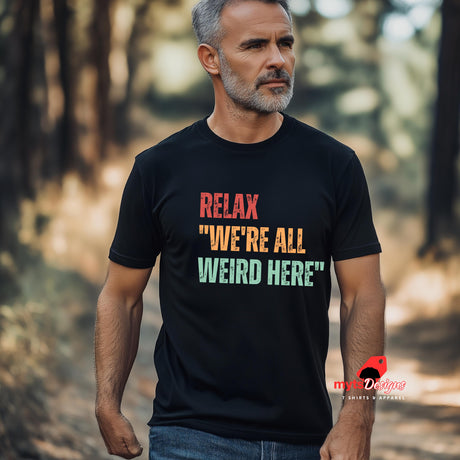 A senior man walking  through the bush wearing a black Retro-style casual tee featuring bold distressed vintage typography reading “Relax — We’re All Weird Here,” printed on ultra-soft Gildan 64000 cotton.