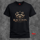 A black short-sleeve crewneck t-shirt with a gold graphic of a raccoon's face and the text 'RACCOON STAY WILD' on the front, displayed against a textured gray background.