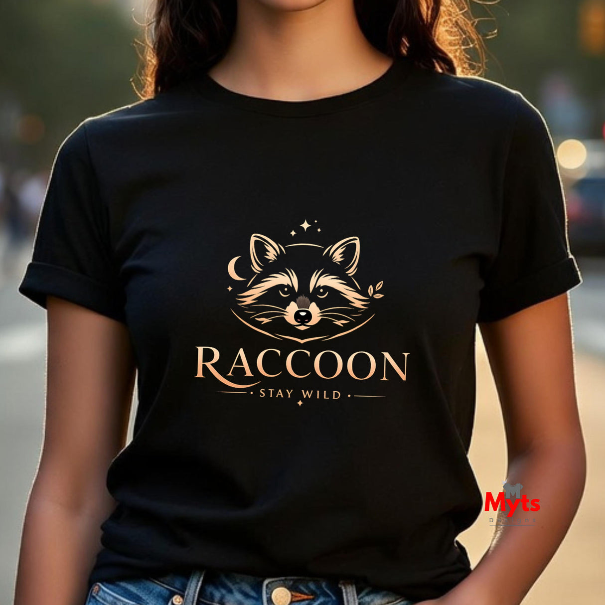 A person wearing a black short-sleeve T-shirt with a gold-toned raccoon graphic and the words 'RACCOON' and 'STAY WILD' on the front, photographed outdoors with a blurred street background.