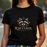A person wearing a black short-sleeve T-shirt with a gold-toned raccoon graphic and the words 'RACCOON' and 'STAY WILD' on the front, photographed outdoors with a blurred street background.