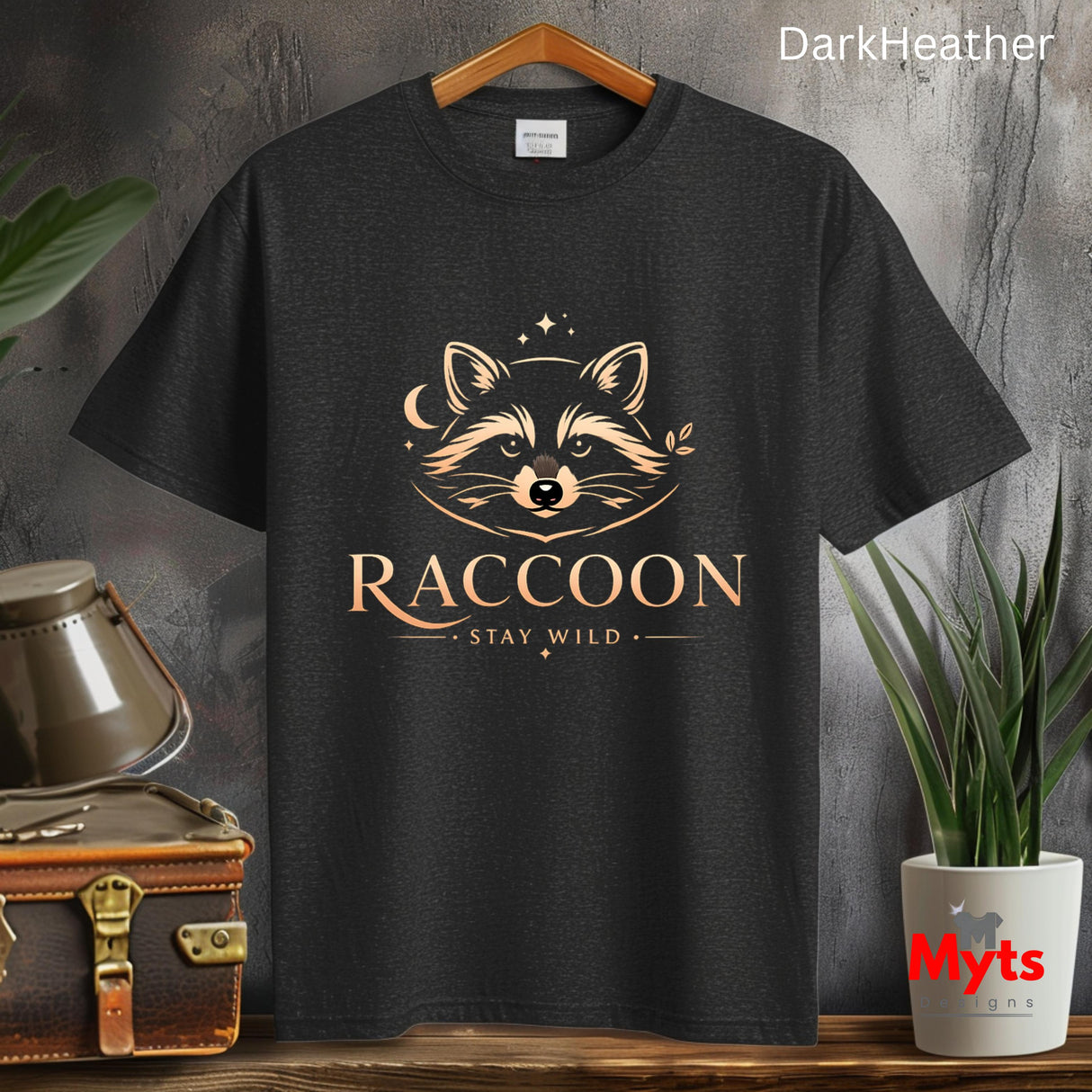 A dark charcoal heather t-shirt hanging on a wooden hanger, featuring a stylized golden-brown raccoon graphic with the text 'RACCOON' and 'STAY WILD'. The background is a rustic gray wall with decorative props including a vintage suitcase, a metal container, and a potted plant.