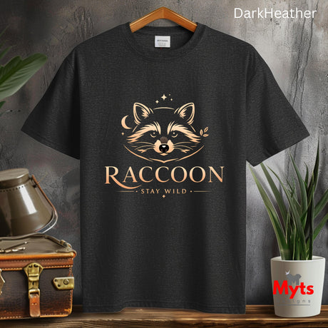A dark charcoal heather t-shirt hanging on a wooden hanger, featuring a stylized golden-brown raccoon graphic with the text 'RACCOON' and 'STAY WILD'. The background is a rustic gray wall with decorative props including a vintage suitcase, a metal container, and a potted plant.