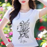 A woman wearing a white t-shirt with a black floral design and 'She Blooms' text, standing in a garden.