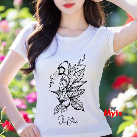A woman wearing a white t-shirt with a black floral design and 'She Blooms' text, standing in a garden.
