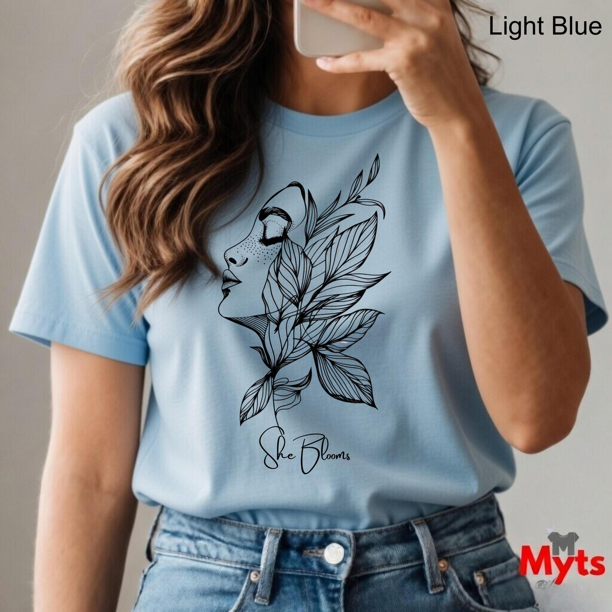 Person wearing a light blue t-shirt with a black floral design and 'She Blooms' text, holding a phone.