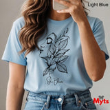 Person wearing a light blue t-shirt with a black floral design and 'She Blooms' text, holding a phone.