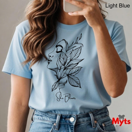 Person wearing a light blue t-shirt with a black floral design and 'She Blooms' text, holding a phone.