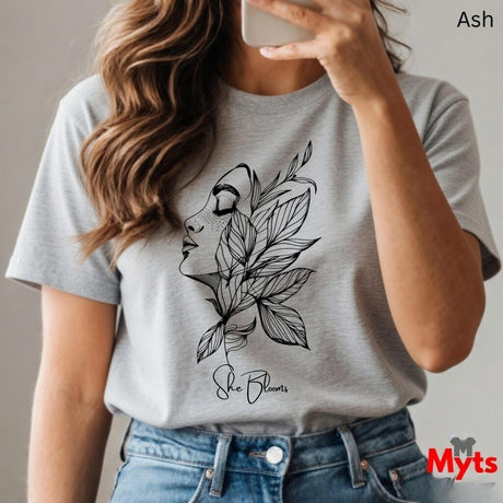 Person wearing a Ash t-shirt with a leaf design and 'She Blooms' text, holding a phone.