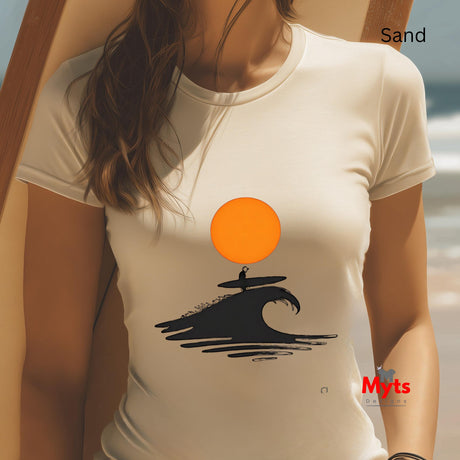 Person wearing a sand colored t-shirt with a graphic design of a surfer on a wave and an orange sun, sitting on sand with blurred ocean background.