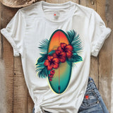 A white T-shirt laid on a wooden surface featuring a vibrant graphic of a surfboard in sunset hues surrounded by red hibiscus flowers and green palm leaves, styled with rolled sleeves and denim shorts.