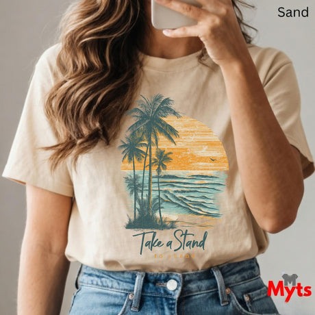 Person wearing a sand t-shirt with a palm tree and sunset design, text 'Take a Stand', and jeans.