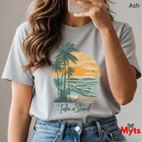 Person wearing a ash t-shirt with a palm tree and sunset design, holding a phone.