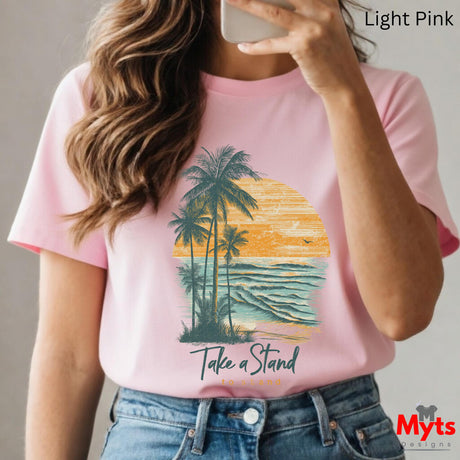 Person wearing a pink t-shirt with a palm tree and sunset design, text 'Take a Stand', and jeans.