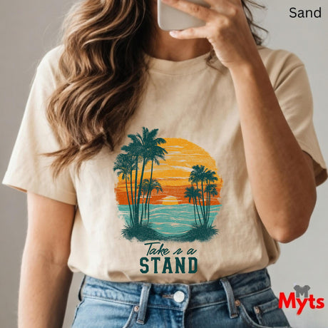 Person wearing a sand t-shirt with a palm tree and sunset design, holding a phone.