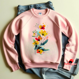 A light pink sweatshirt featuring an artistic watercolor floral design with red, yellow, and orange wildflowers accented by lush green leaves and delicate splatters.

t