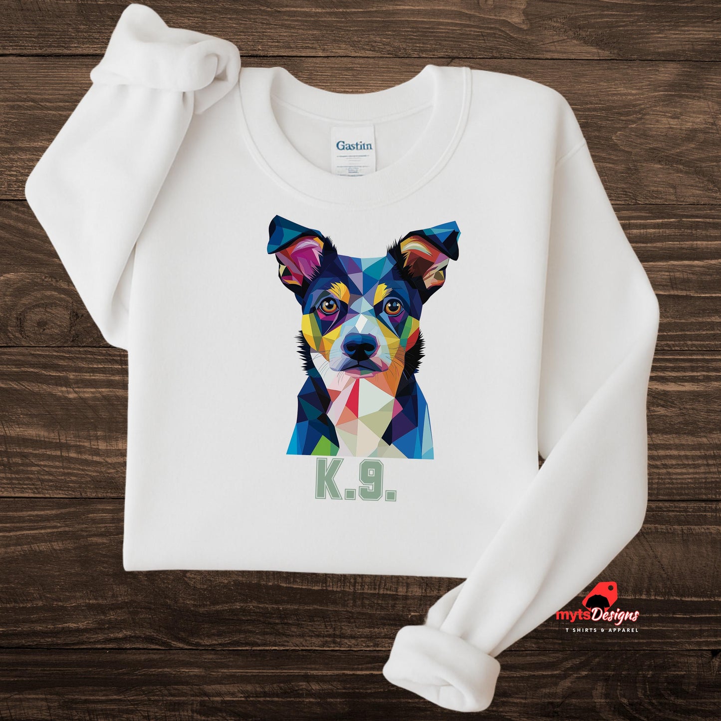 A white sweatshirt displayed on a wooden surface featuring a bold and colorful polygon art dog portrait with the text “K.9.” beneath it, designed by MytsDesigns.

