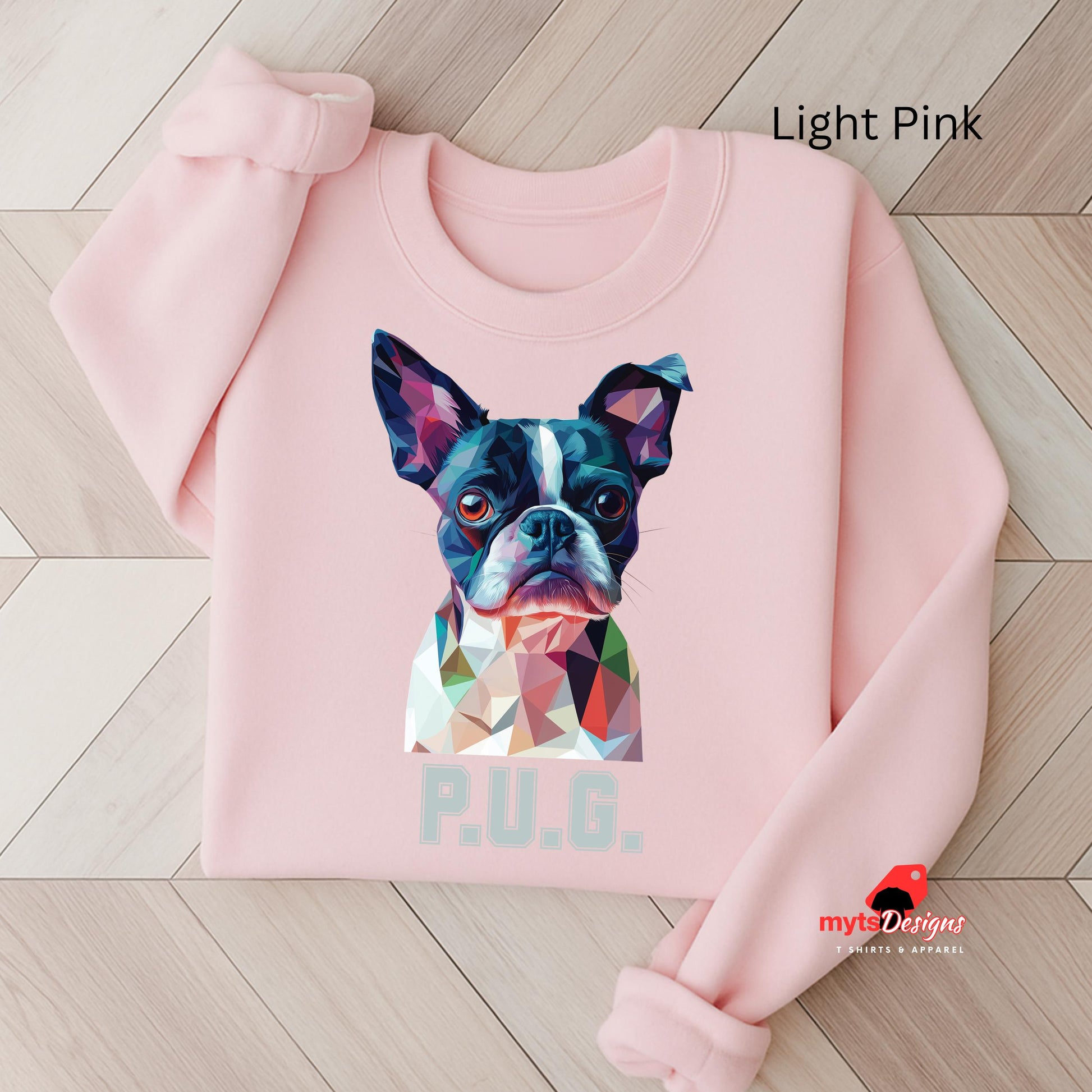 A Light Pink sweatshirt displayed on a wooden surface featuring a vibrant polygon-style dog portrait with the text “P.U.G.” below, designed by MytsDesigns