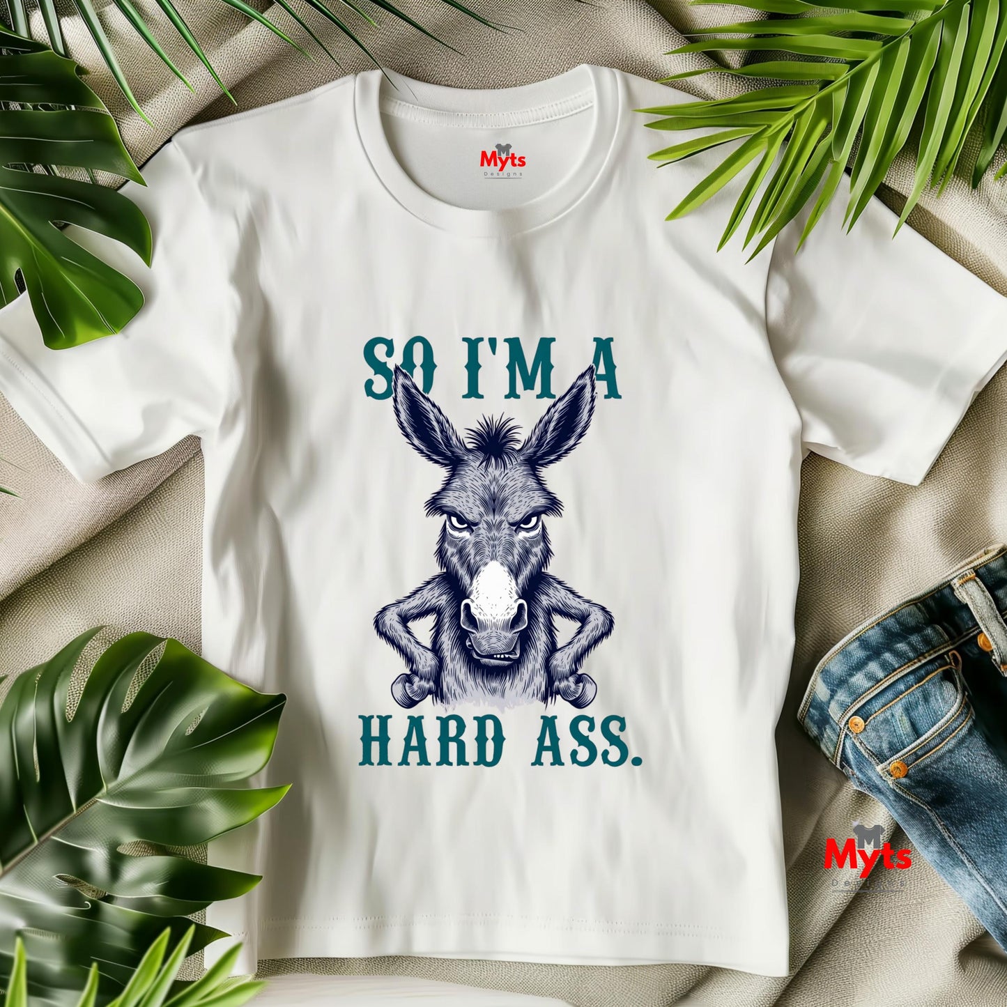 White t-shirt with a donkey graphic and text, surrounded by green leaves on a beige surface.