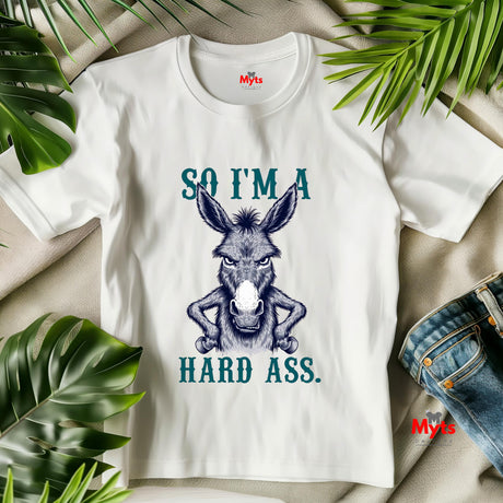 White t-shirt with a donkey graphic and text, surrounded by green leaves on a beige surface.