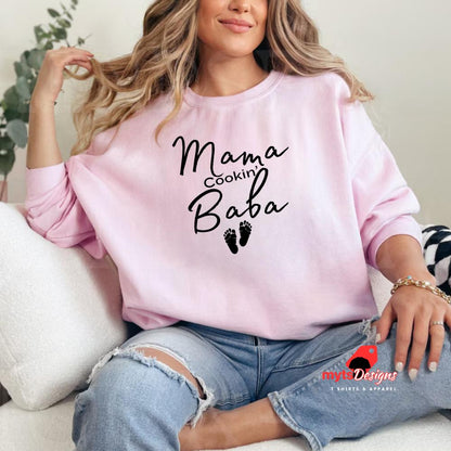 A woman lounging in a pink sweatshirt with black text that reads “Mama Cookin’ Baba” above two small baby footprints, styled for maternity comfort.