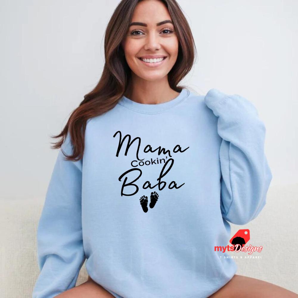 A woman lounging in a light blue sweatshirt with black text that reads “Mama Cookin’ Baba” above two small baby footprints, styled for maternity comfort.