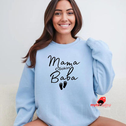 A woman lounging in a light blue sweatshirt with black text that reads “Mama Cookin’ Baba” above two small baby footprints, styled for maternity comfort.