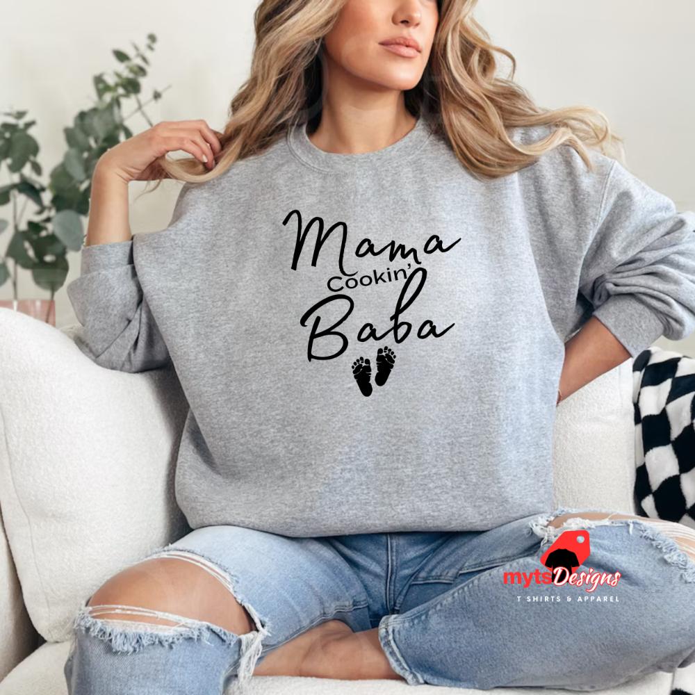 A woman lounging in a sports grey sweatshirt with black text that reads “Mama Cookin’ Baba” above two small baby footprints, styled for maternity comfort.