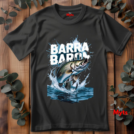 Black t-shirt laid flat on wooden surface with “BARRA BARON” text and large realistic jumping barramundi fish graphic splashing out of blue water with fishing lure